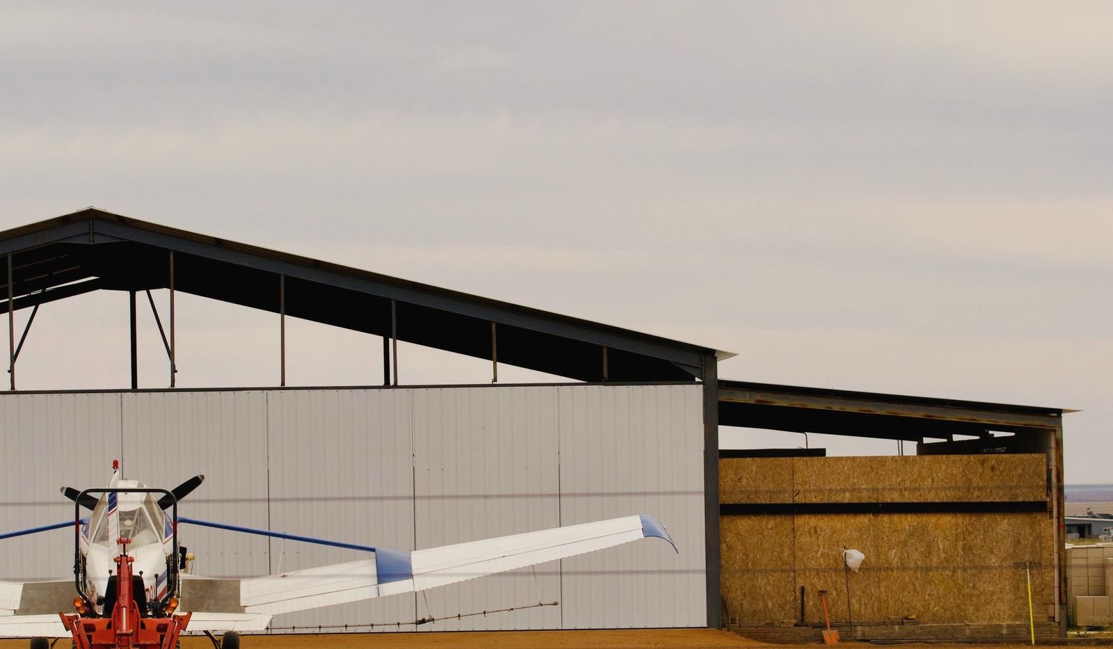 An open steel hangar on flat dry ground with an agricultural airplane and tug outside