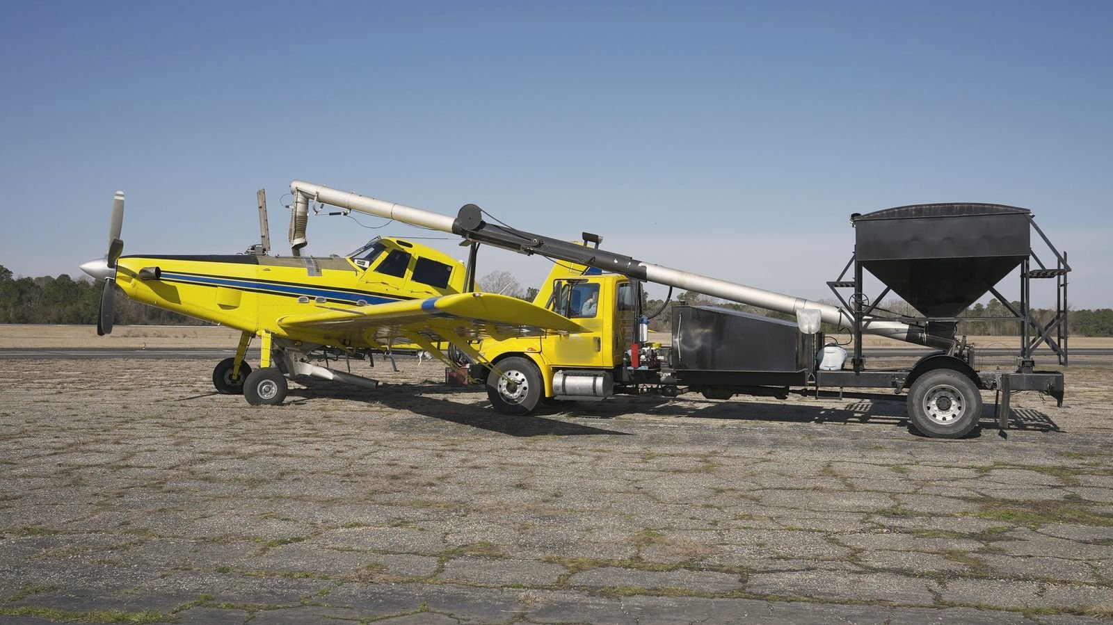 A yellow agricultural airplane loading from a tender truck on a cracked ramp under a clear sky